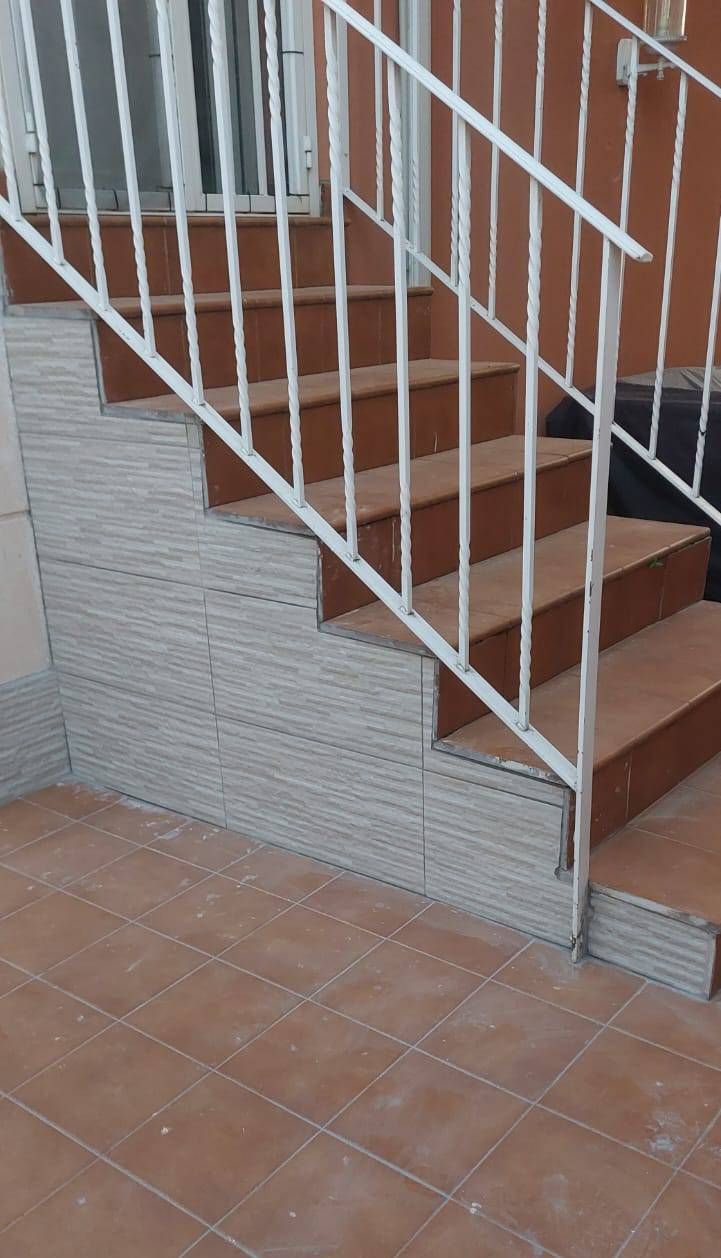 Escalera con barandilla blanca, paneles de yeso apilados en el lateral, escalones marrones, suelo de terracota, ambiente interior.