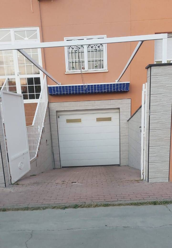 Edificio naranja con puerta de garaje blanca y rampa de entrada. Toldo azul sobre el garaje y valla blanca.