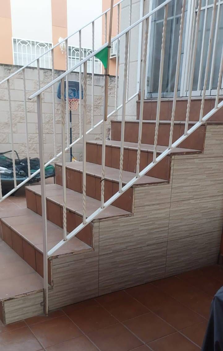 Escalera exterior con barandilla metálica, peldaños marrones, paredes de azulejos beige.