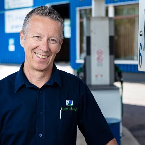 Hymyilevä mies tummansinisessä paidassa seisoo sisätiloissa sinivalkoisen sisäänkäynnin lähellä