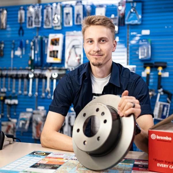 Mekaanikko pitelee jarrulevyä autovaraosaliikkeessä, ja hänen takanaan sinisellä seinällä on esillä työkaluja.