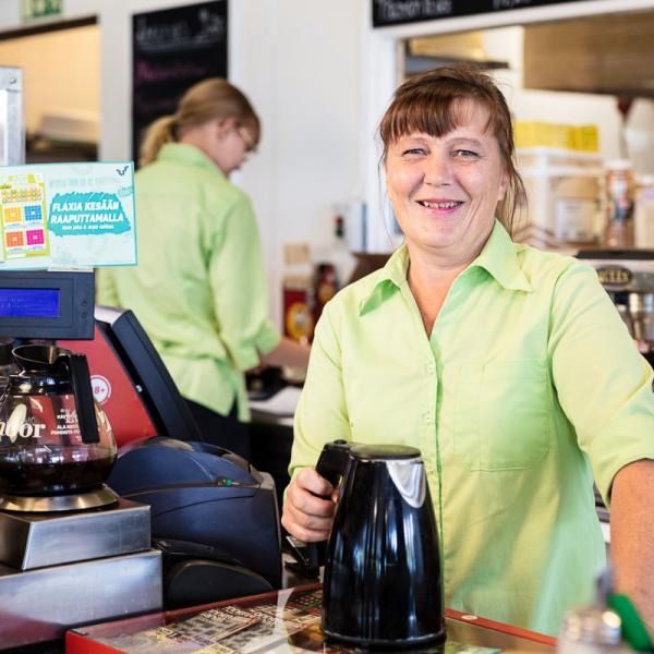 Vaaleanvihreään paitaan pukeutunut barista hymyilee kahvilan tiskin takana musta vedenkeitin kädessään.