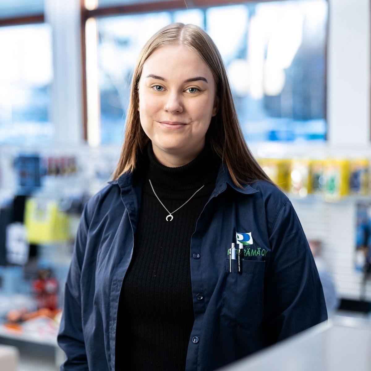 Hymyilevä työntekijä tummansinisessä paidassa ja takissa seisoo valoisalla kaupan käytävällä