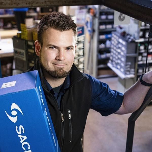 Mies kaupassa pitelee sinistä laatikkoa hyllyjen lähellä ja katsoo kameraan