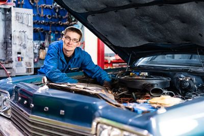 Siniseen takkiin pukeutunut mekaanikko työskentelee klassikkoauton parissa autotallissa konepelti auki