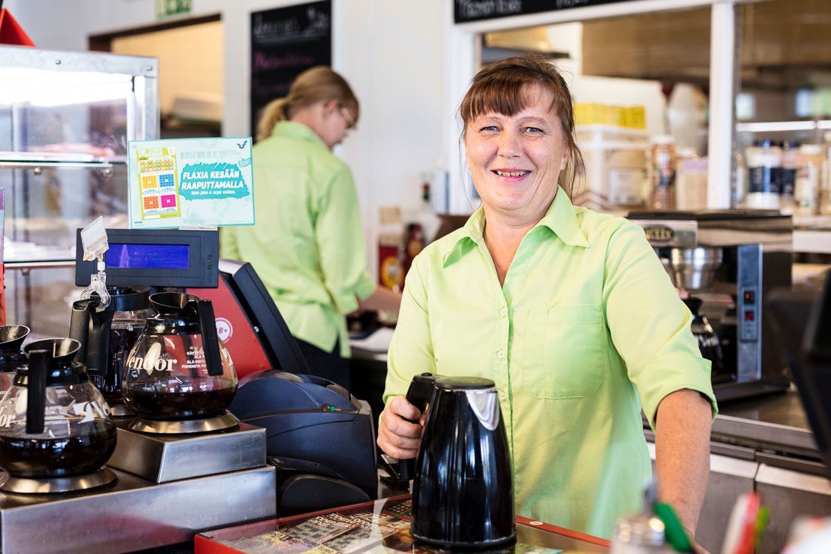Smiling café worker in a lime-green shirt behind the counter with coffee equipment and a kettle