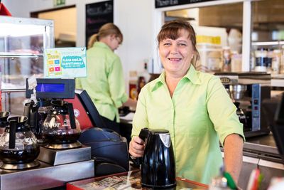 Vihreäpaitainen barista hymyilee kahvilan tiskin takana ja pitää kädessään mustaa vedenkeitintä