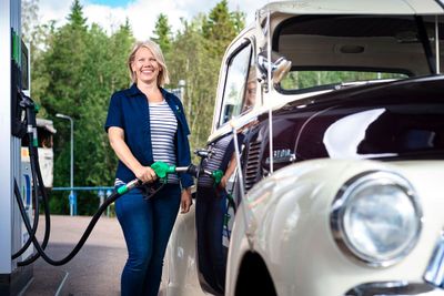 Nainen tankkaa valkoista vintage-autoa ulkoilmahuoltoasemalla hymyillen metsäisessä ympäristössä