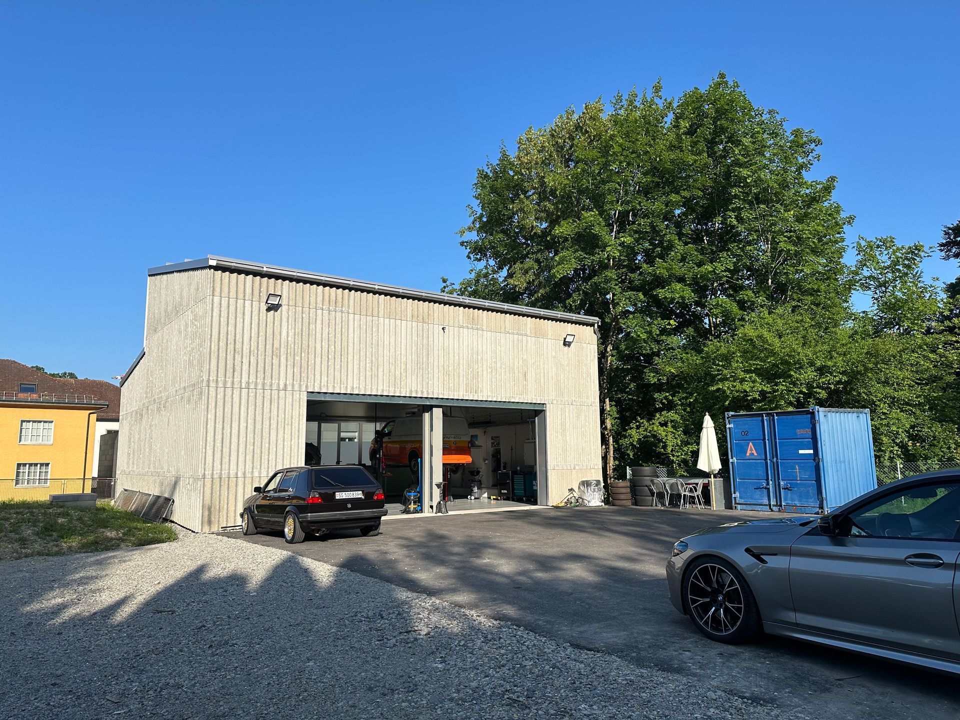 Garage with open doors, a black car parked inside, and a gray car outside on a gravel lot.