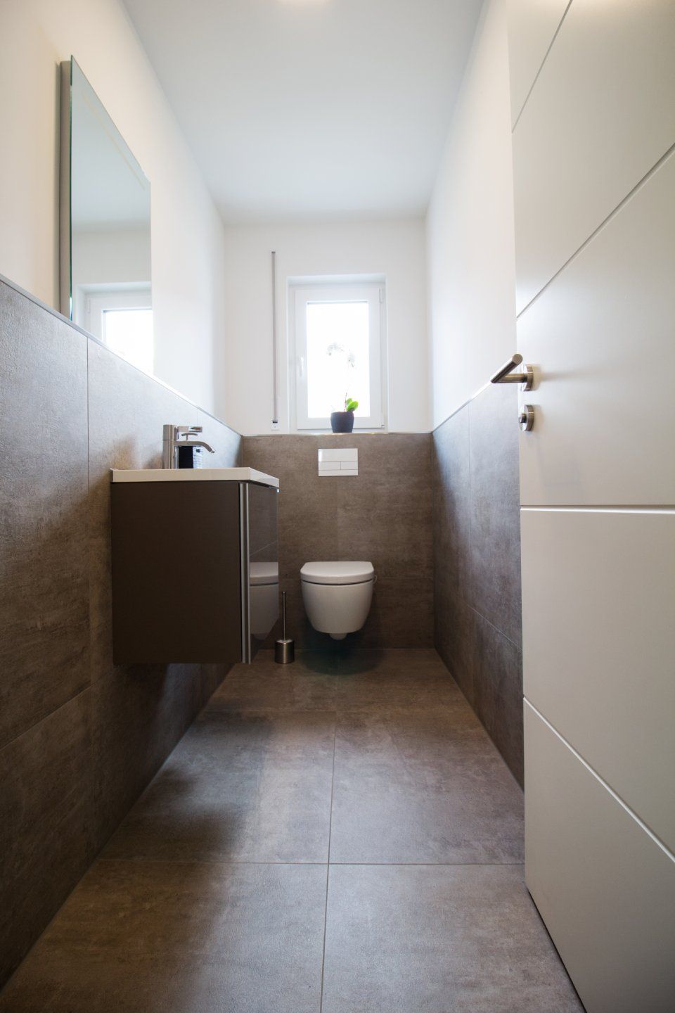 Schmale, moderne Badezimmer mit gefliesten Wänden, wandhängendem WC, Waschbecken, Spiegel und Fenster am Ende.