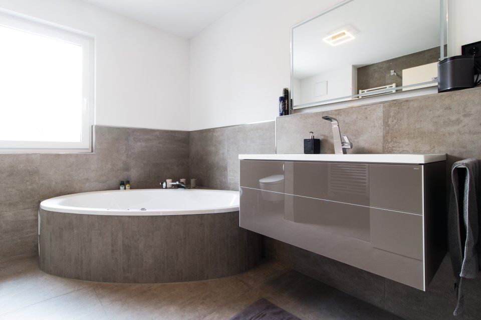 Modernes Badezimmer mit freistehender ovaler Badewanne, schwebendem Waschtisch, großem Spiegel und grauen Fliesen an Wänden und Boden.
