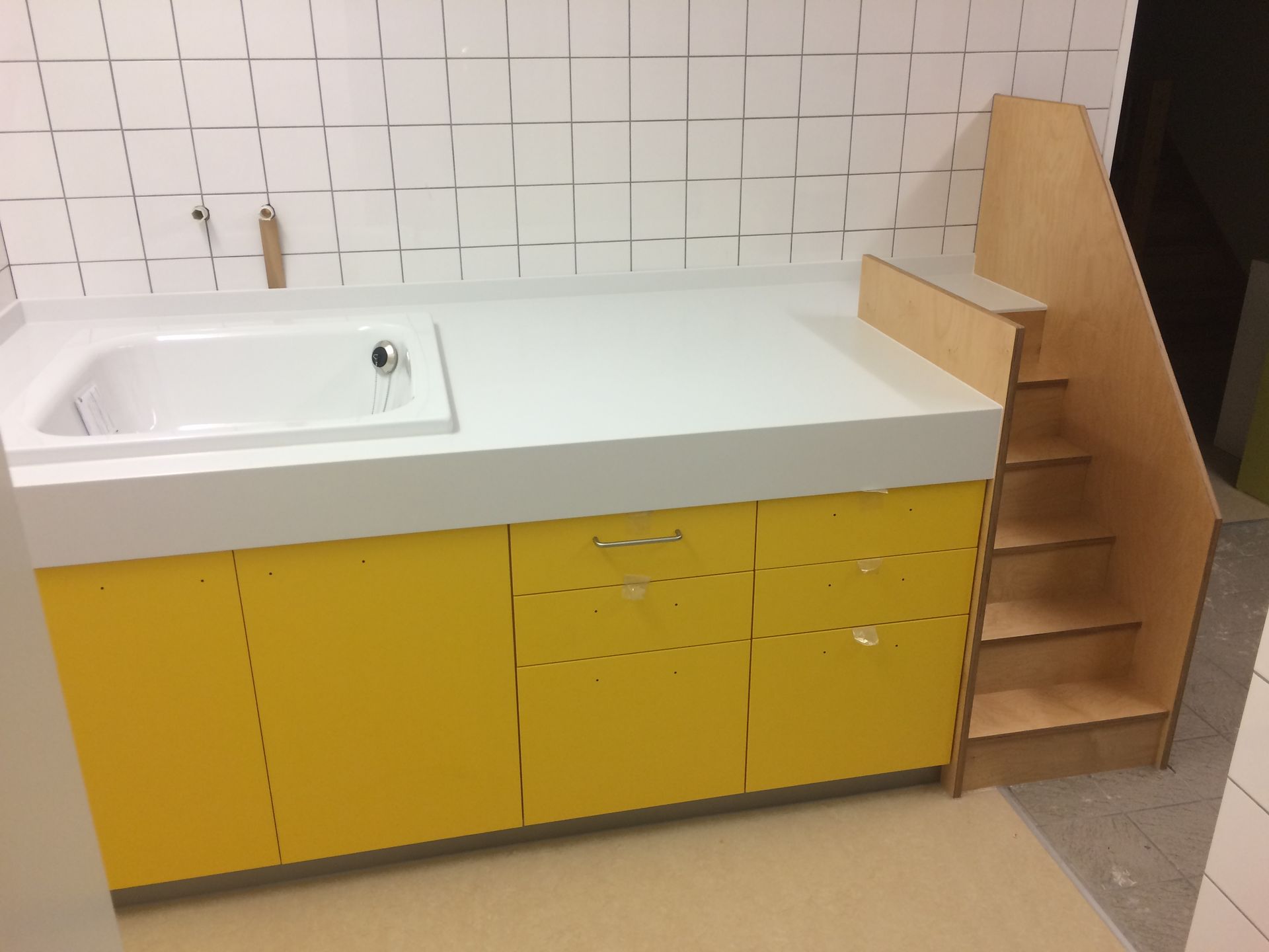 Wickelkommode im Kindergarten, mit Duschtasse und Holztreppe, gelbe Fronten, Oberfläche Weiß