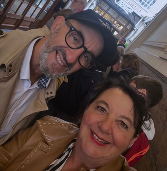 Un couple souriant prend un selfie en extérieur, portant des vestes, des lunettes et un chapeau ; une scène de rue en arrière-plan.