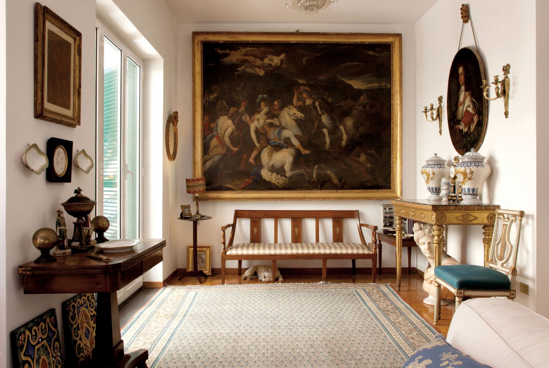 Pièce meublée d'antiquités, avec un grand tableau, une fenêtre et des objets décoratifs sur les tables.