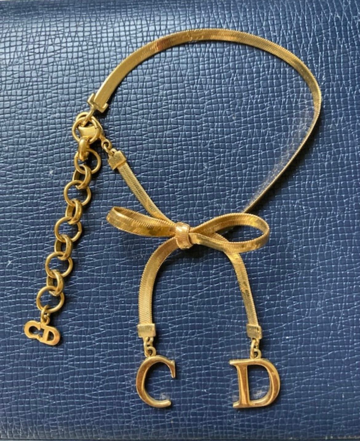 Collier Dior doré orné d'un nœud, avec des breloques CD pendantes, posées sur une surface bleue texturée.