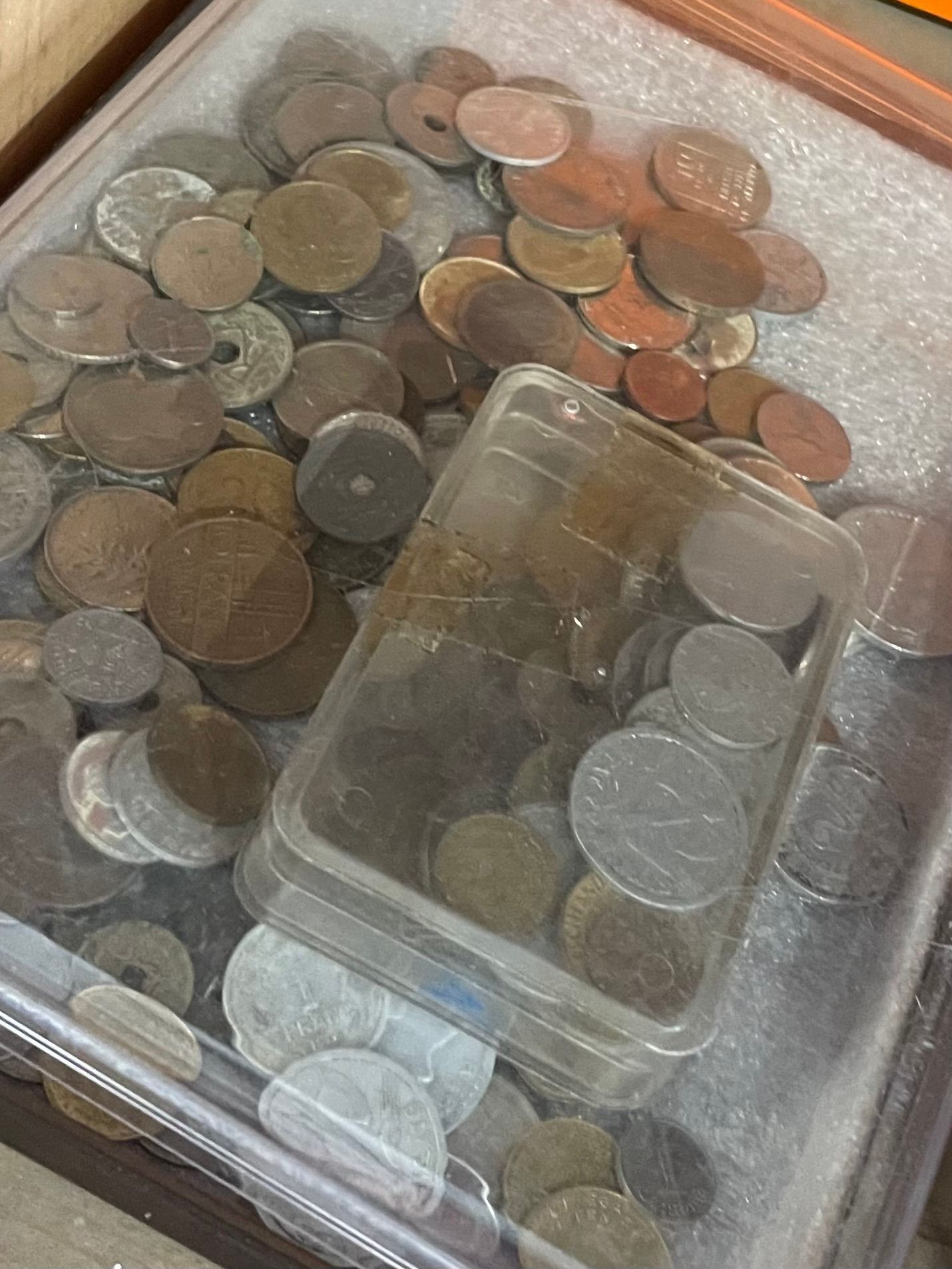 Pièces de monnaie anciennes assorties dans un récipient en plastique transparent et ses alentours. Tailles et couleurs variées.