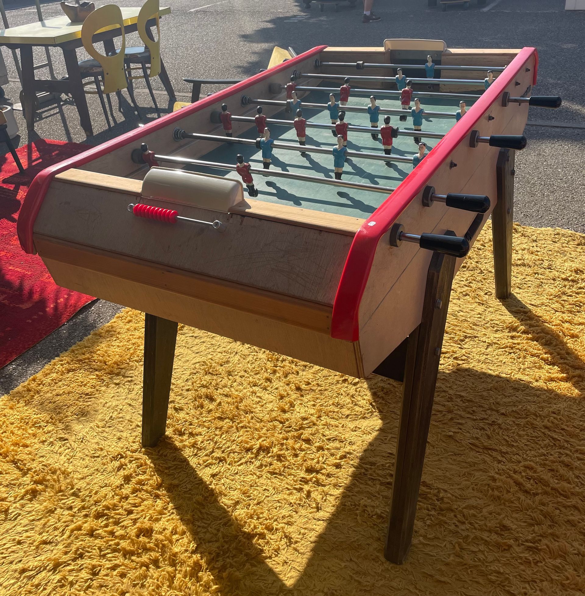 Baby-foot sur un tapis jaune à l'extérieur, joueurs rouges et bleus, bordures rouges.