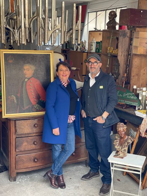 Deux personnes posent avec un tableau ancien et des meubles dans une boutique encombrée.