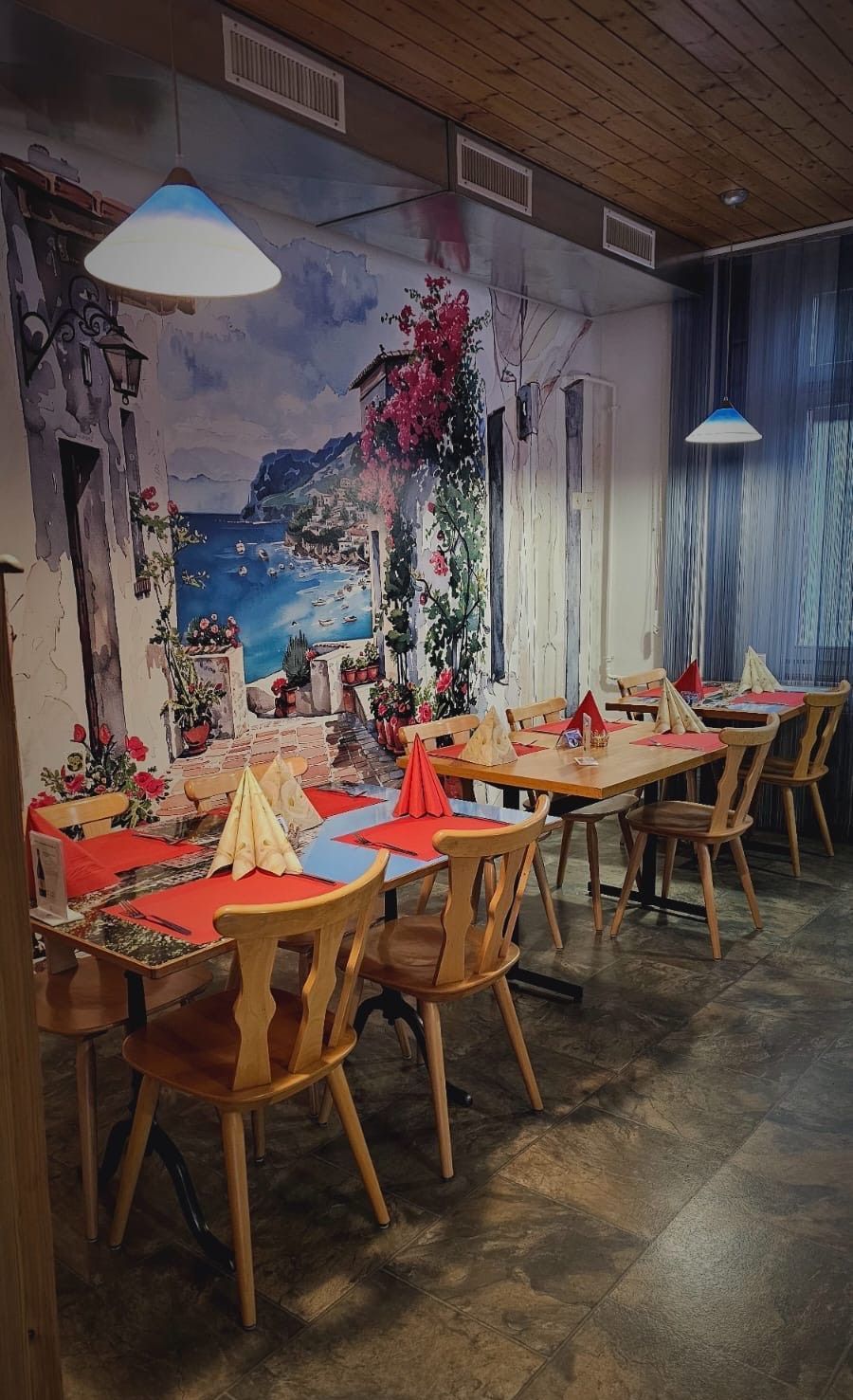 salle de restaurant avec des tables, des chaises, des sets rouges et des fresques de paysage de mer sur les murs