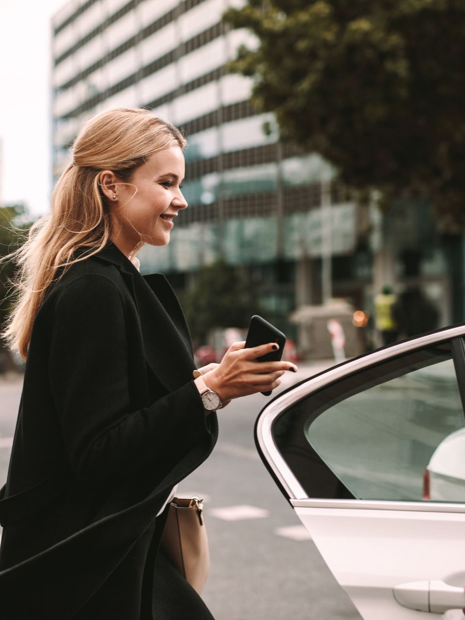 Une femme blonde en manteau noir utilise un téléphone, sur le point d'entrer dans une voiture blanche dans la rue de la ville.