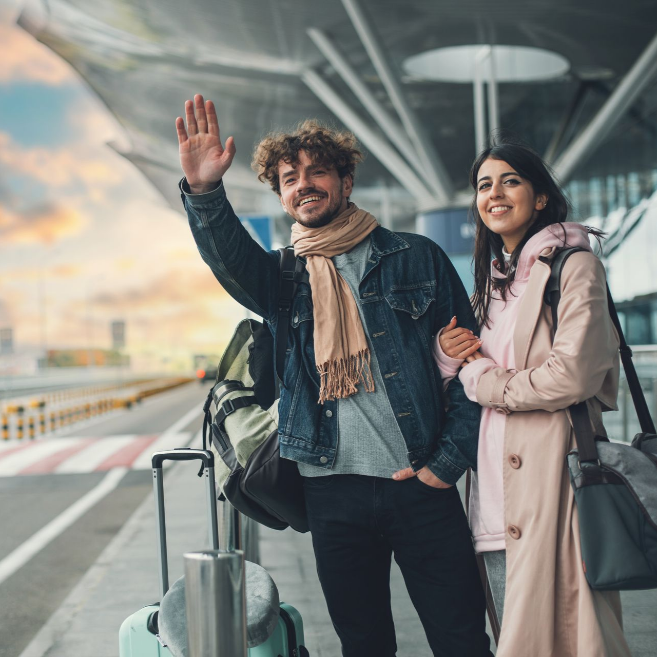 Un couple à l'aéroport. L'homme salue, la femme sourit. Ils ont des bagages et portent des vestes.