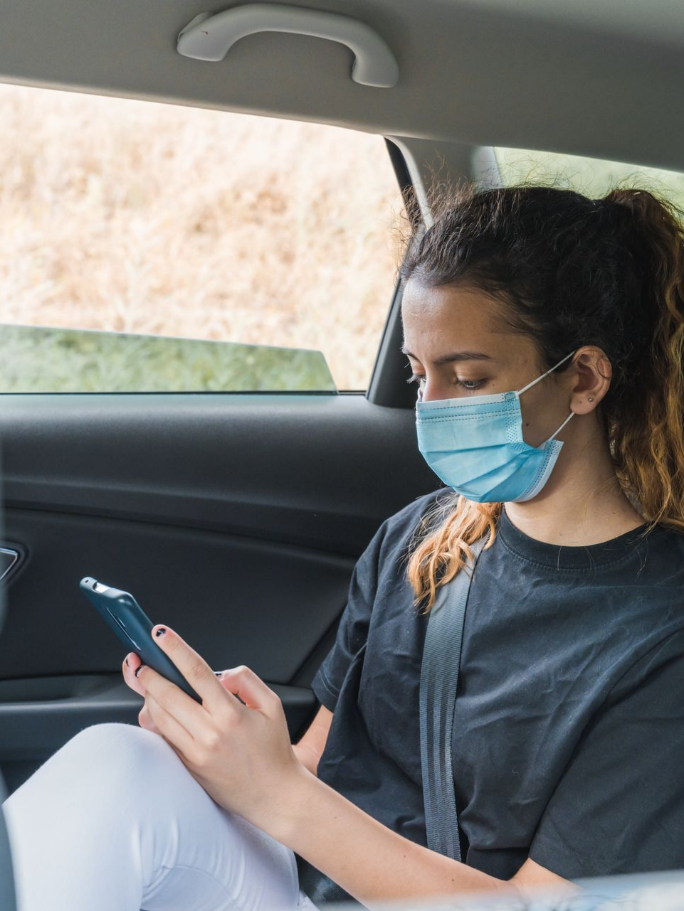 Une femme dans une voiture portant un masque facial, regardant son téléphone