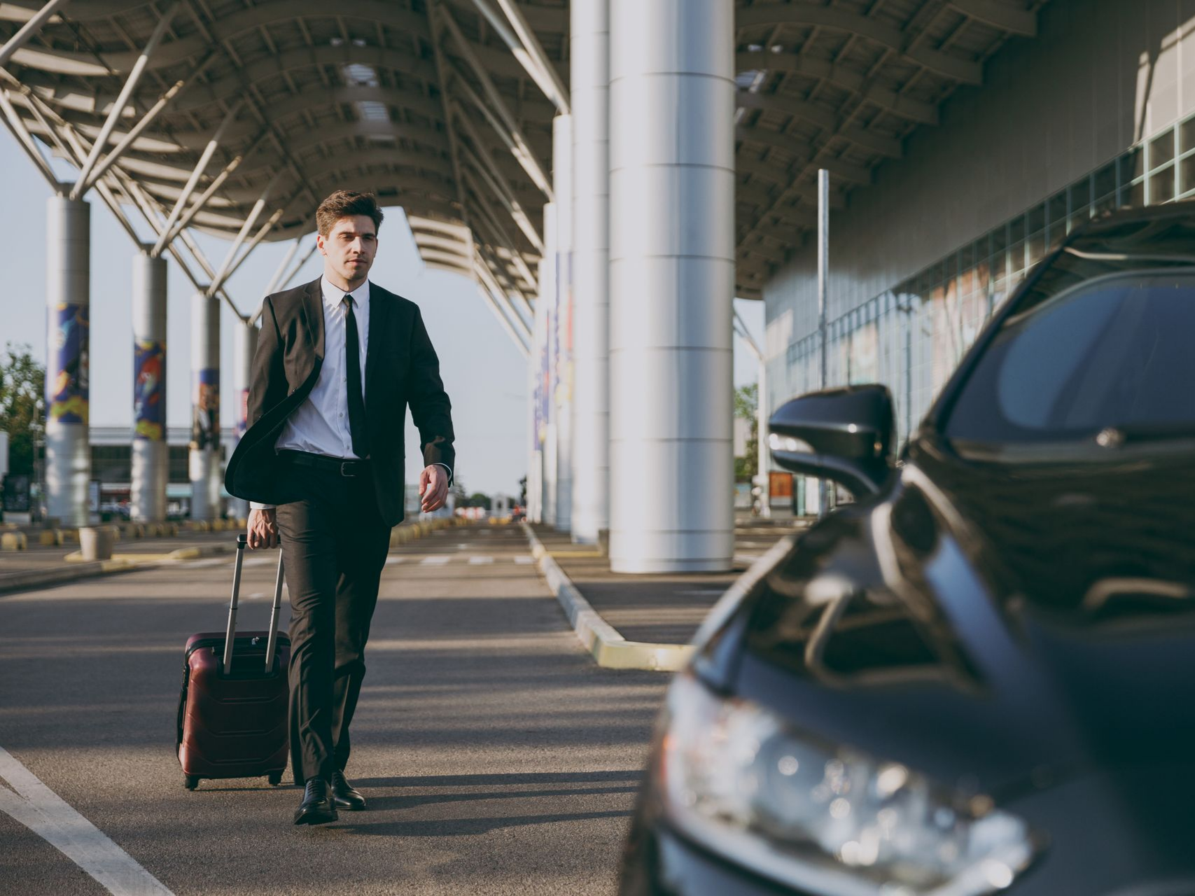 Un homme en costume marchant avec des bagages vers une voiture noire qui l'attend dans un aéroport.