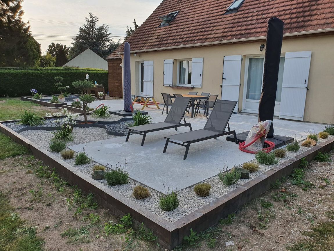 Terrasse avec chaises longues, parasol et table devant une maison. Des parterres de fleurs bordent la terrasse.