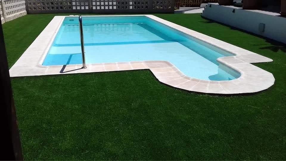 Piscina al aire libre con agua azul cristalina, terraza blanca y césped artificial verde a su alrededor.