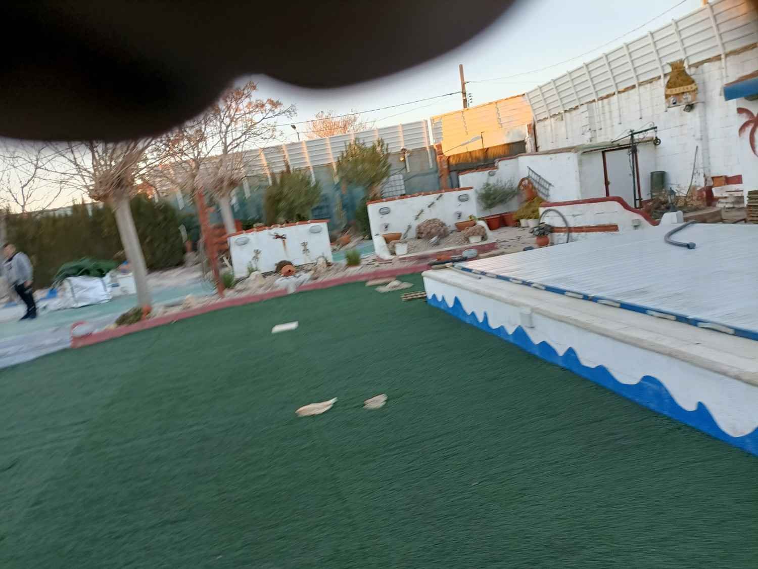 Campo de minigolf al aire libre con césped verde, paredes blancas y una persona caminando al fondo.