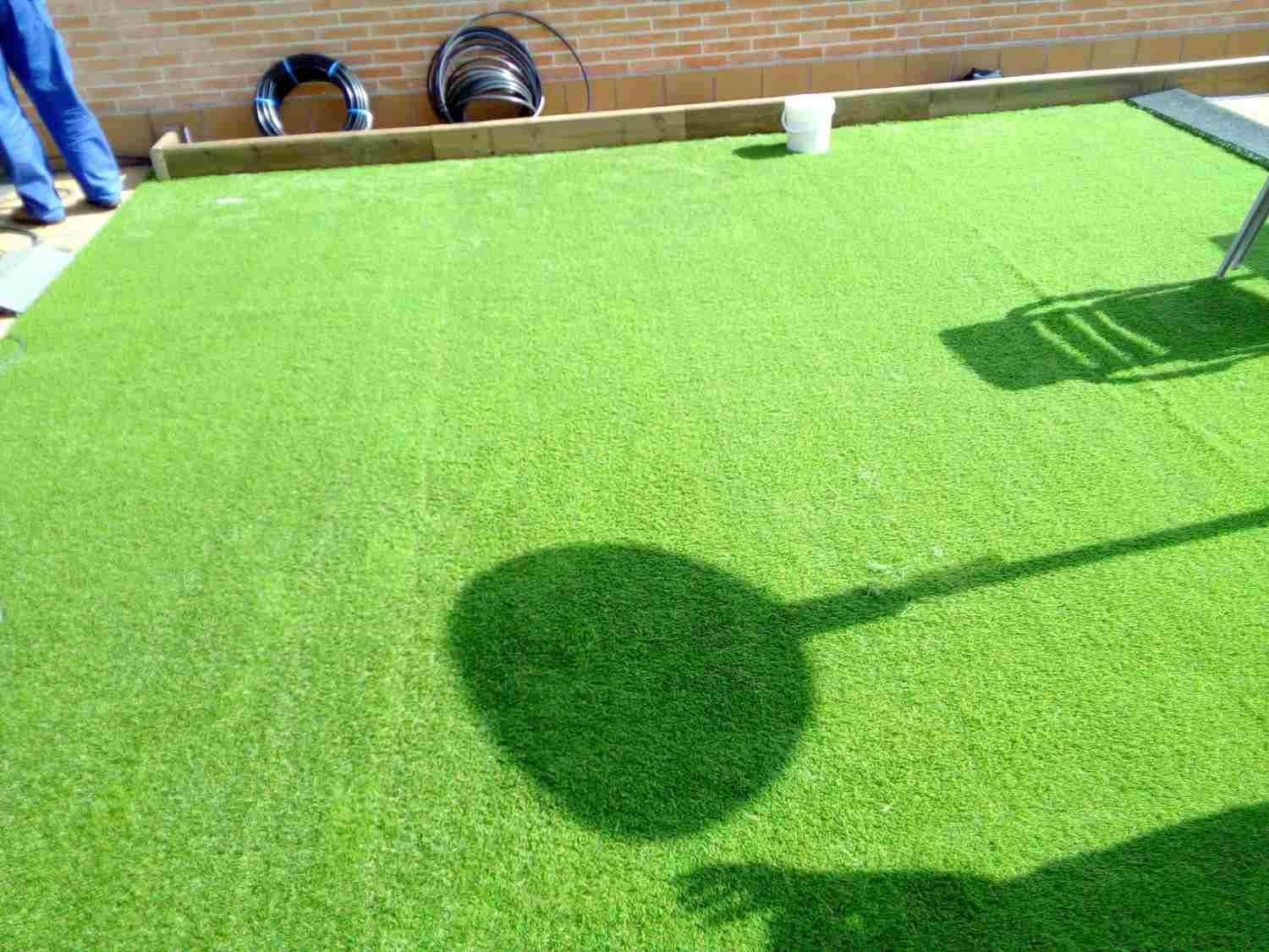 Campo de minigolf con la sombra de un palo de golf y una pelota proyectada sobre el césped cerca de una pared de ladrillos.