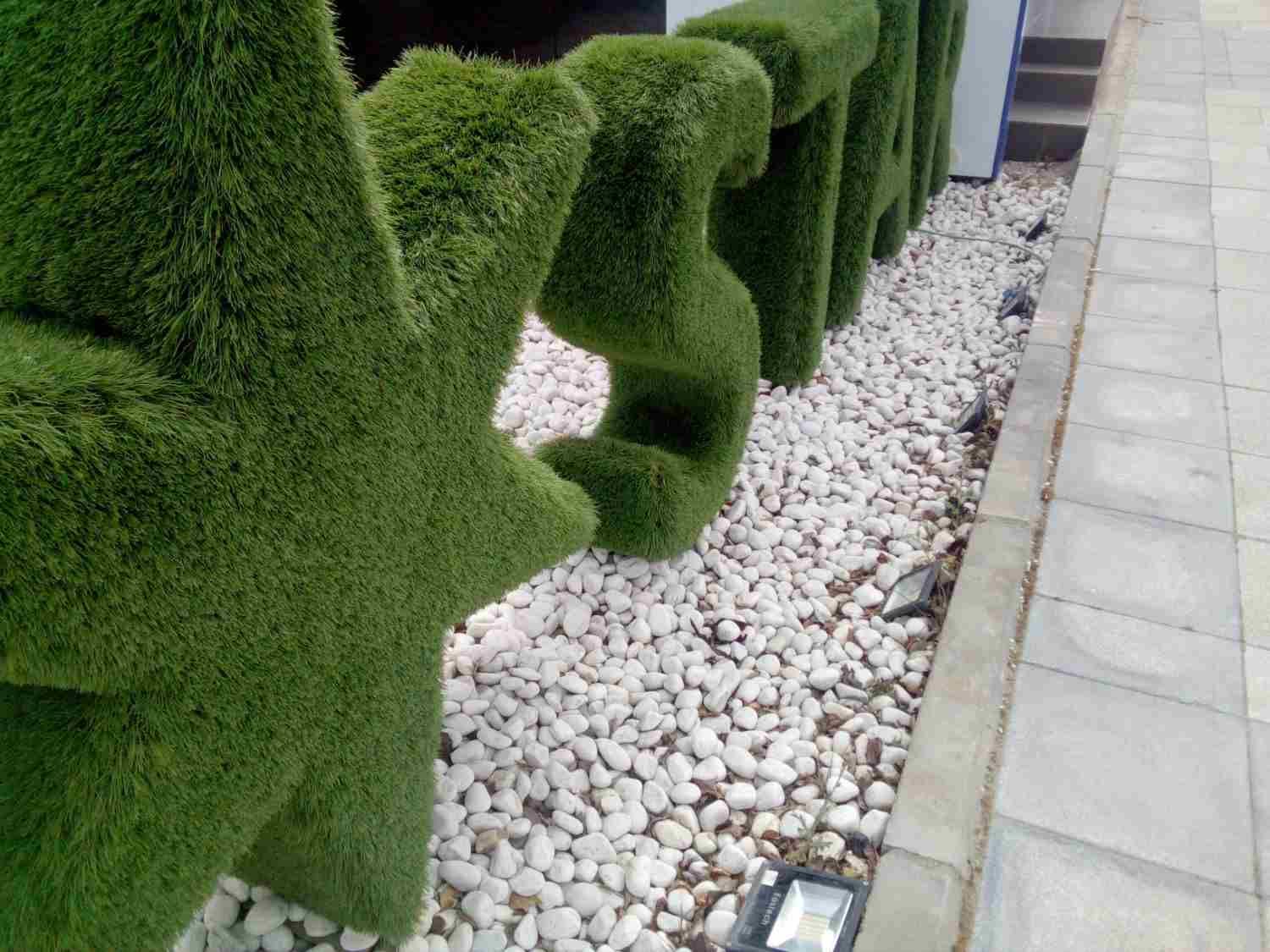 Un seto ornamental con forma de letras se encuentra junto a una franja de grava blanca y un sendero pavimentado.