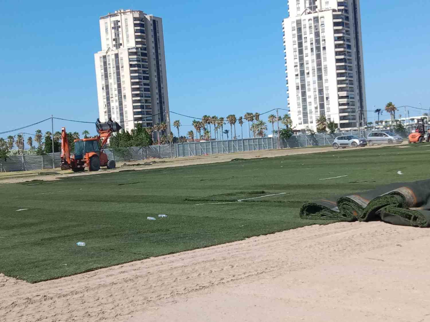 Campo deportivo verde con un tractor, arena en primer plano y dos torres altas al fondo.