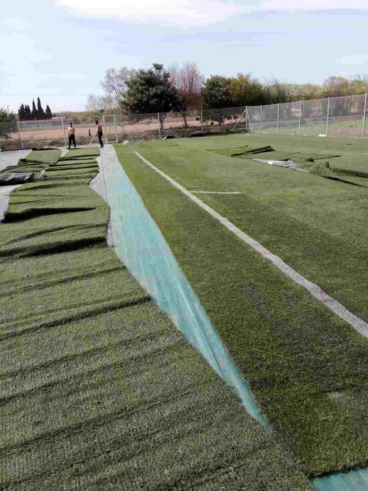 Campo de césped artificial con una larga línea divisoria azul y algunas personas a lo lejos bajo un cielo nublado.