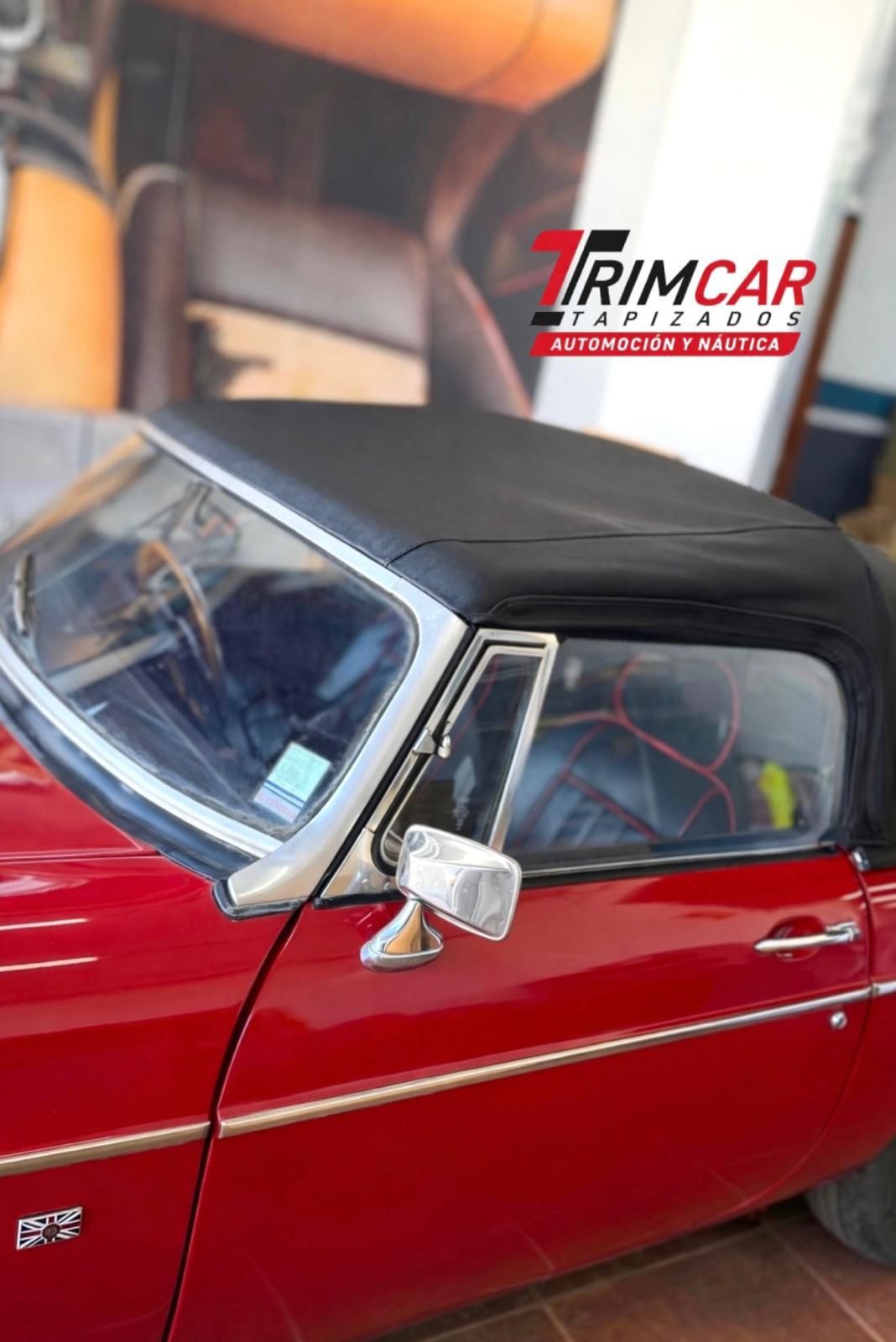 Coche clásico descapotable rojo con capota negra aparcado cerca de un cartel de Trimcar.
