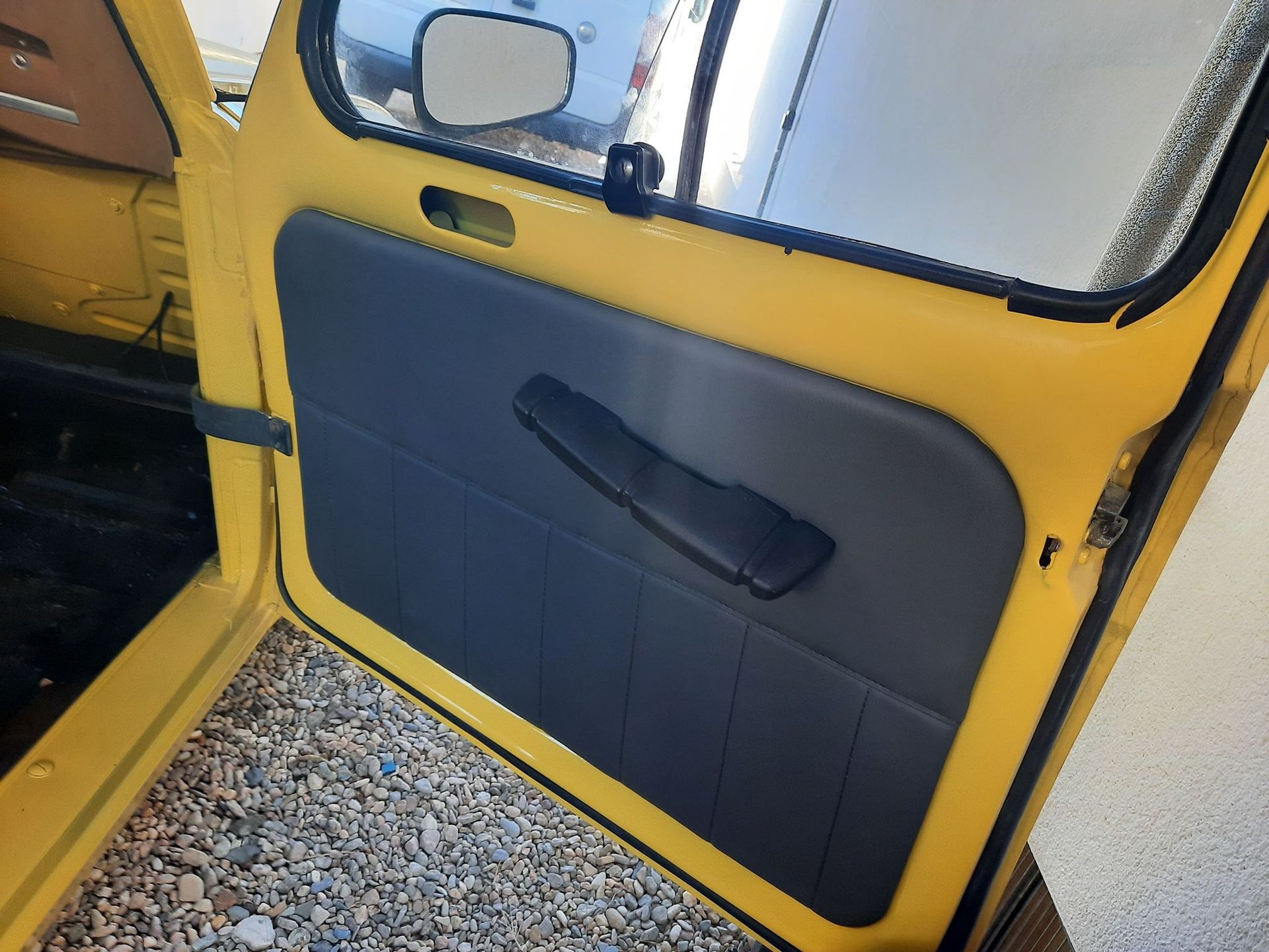 Primer plano de la puerta de un vehículo amarillo con un panel azul oscuro y manija negra, abierta hacia un suelo de grava.