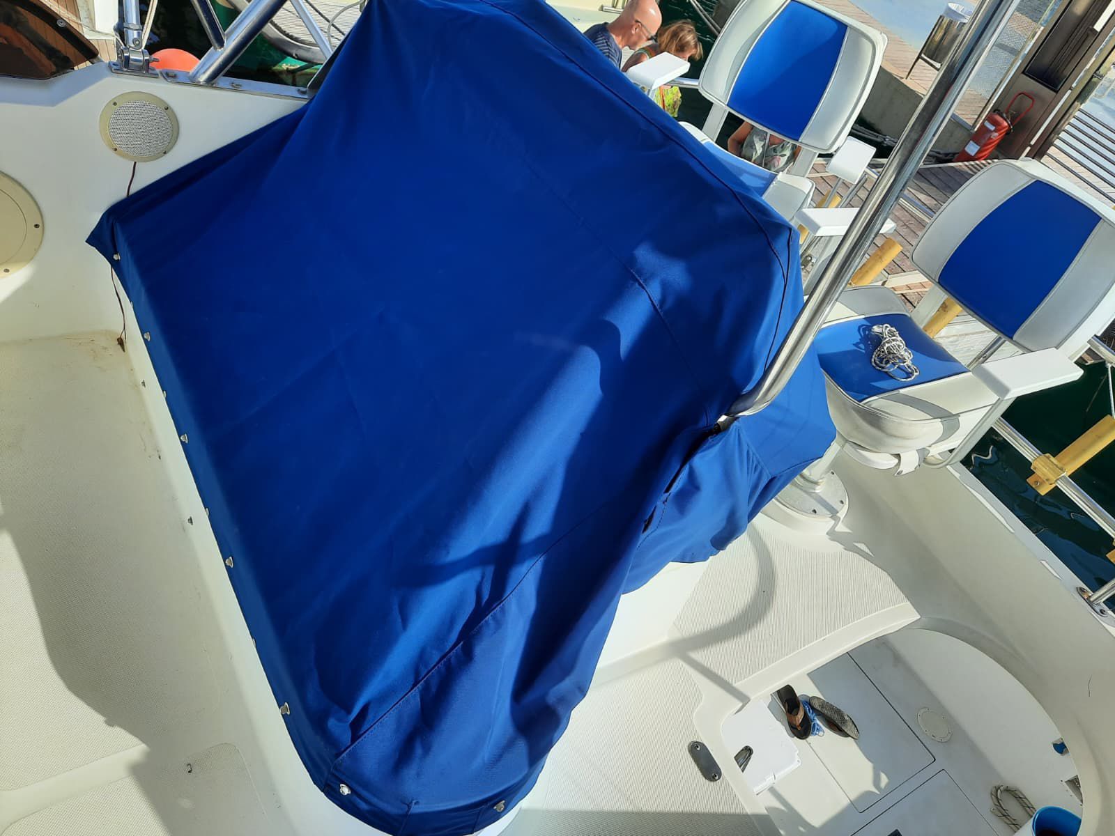 Asiento de barco tapizado de azul sobre una cubierta blanca con barandillas a su alrededor y agua al fondo.