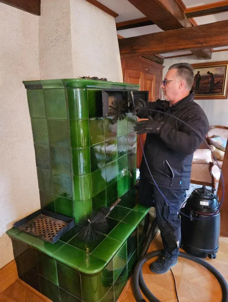 Un professionnel ramoneur nettoie un poêle en maçonnerie carrelé de vert à l'aide d'une brosse et d'un aspirateur dans une pièce.