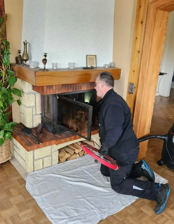 Un ramoneur est agenouillé sur un tapis blanc, en train de nettoyer un insert de cheminée dans un salon.