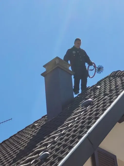 Un ramoneur se tient sur un toit de tuiles tenant une brosse de ramonage, à côté d'une cheminée.