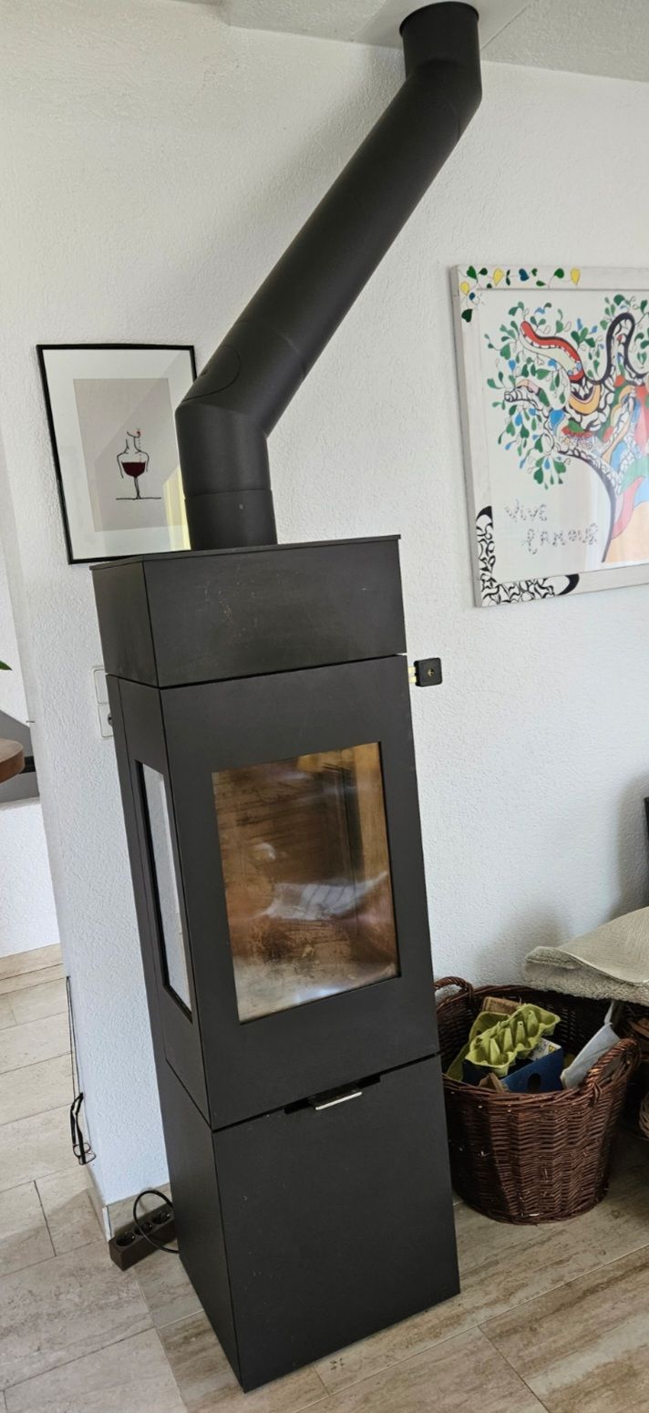 Poêle à bois noir sur pied avec conduit de cheminée incliné dans une pièce, à côté d'un tableau encadré et d'un panier.