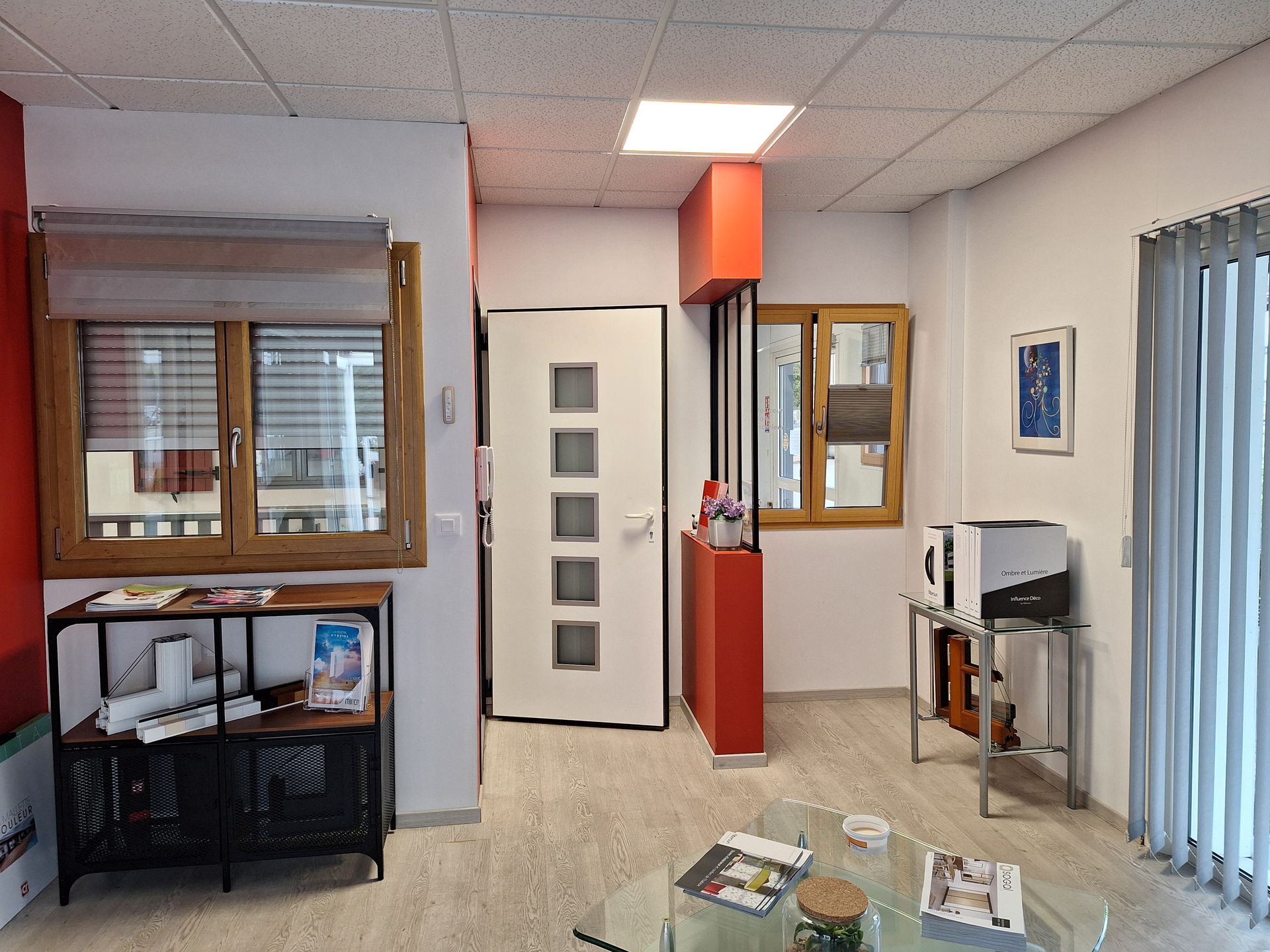 Une salle d'attente avec parquet clair, une porte blanche à cinq fenêtres, une cloison latérale orange et des tables en verre.