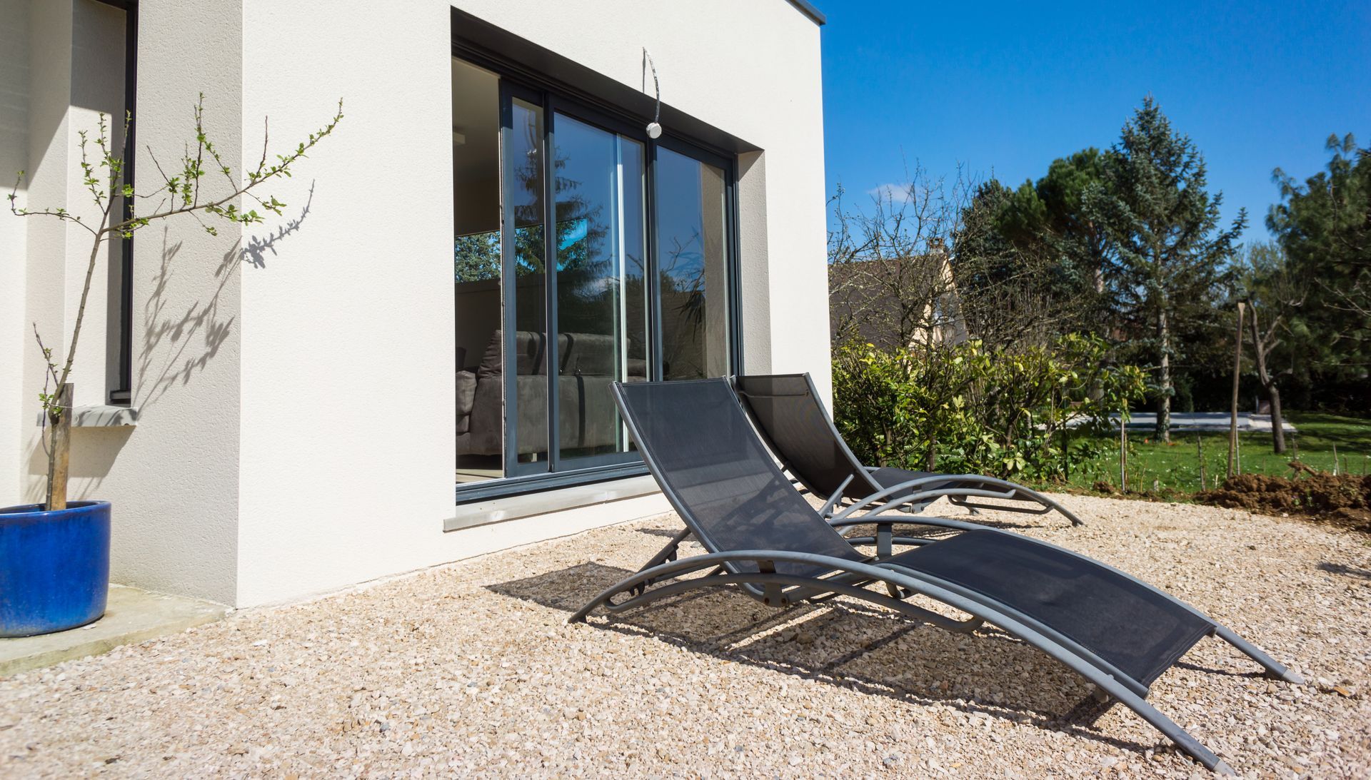 Deux chaises longues noires sur une terrasse en gravier devant une maison moderne avec des portes coulissantes en verre et une jardinière bleue à proximité.