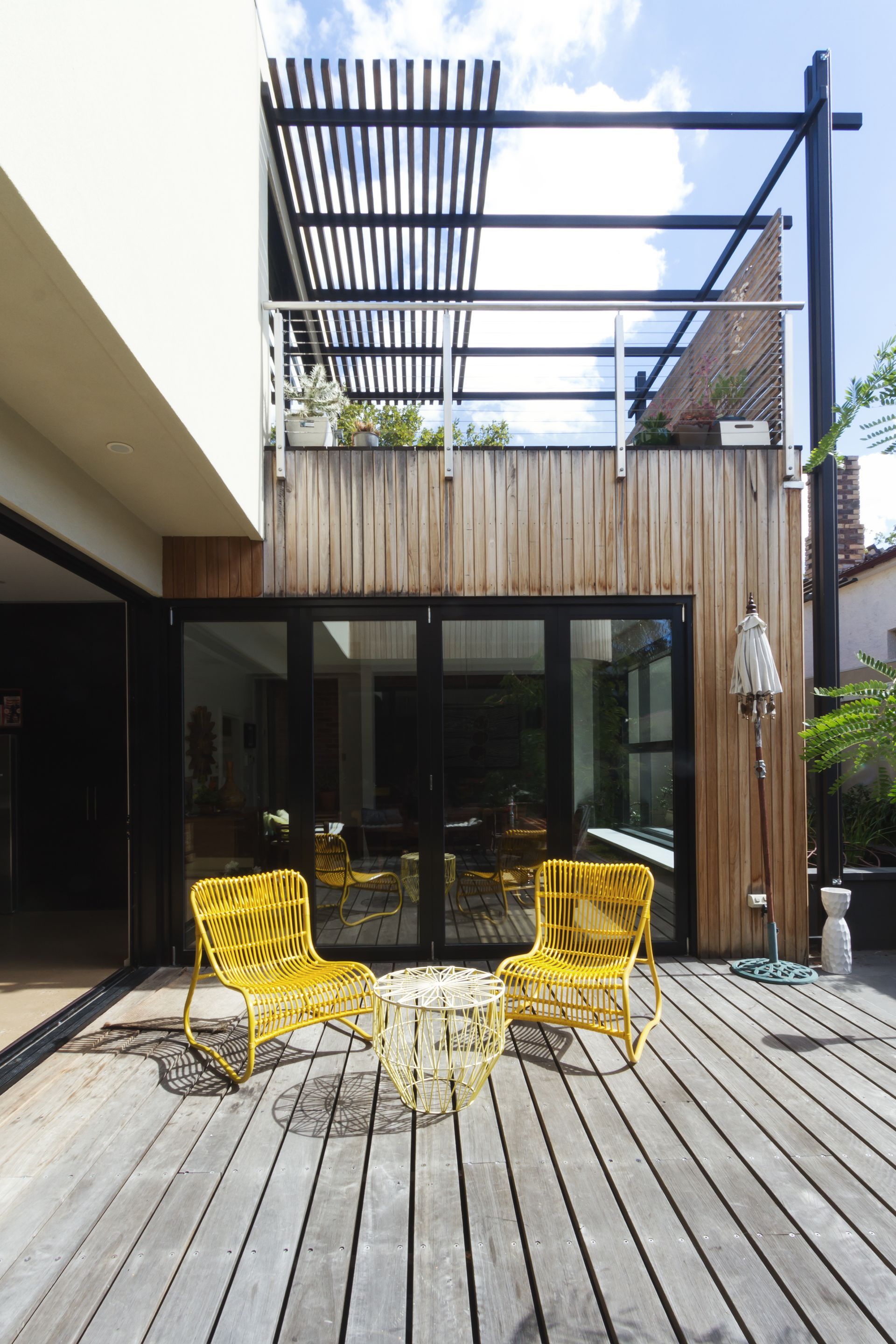 Deux chaises de patio jaunes et une petite table assortie sont installées sur une terrasse en bois devant une maison moderne aux portes vitrées noires.