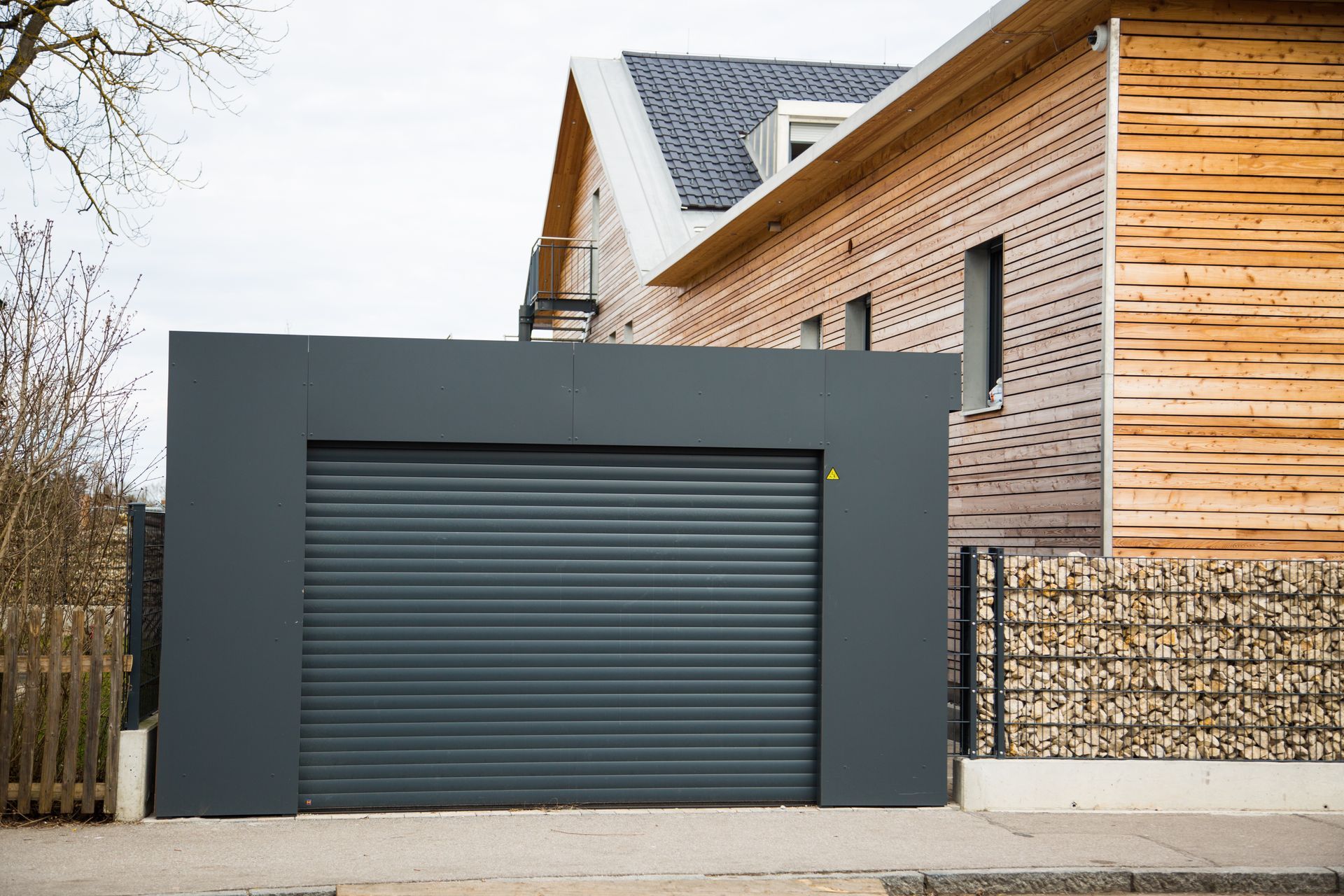 Un garage gris anthracite avec une porte coulissante jouxte une maison au bardage en lattes de bois et au mur en gabions de pierre.