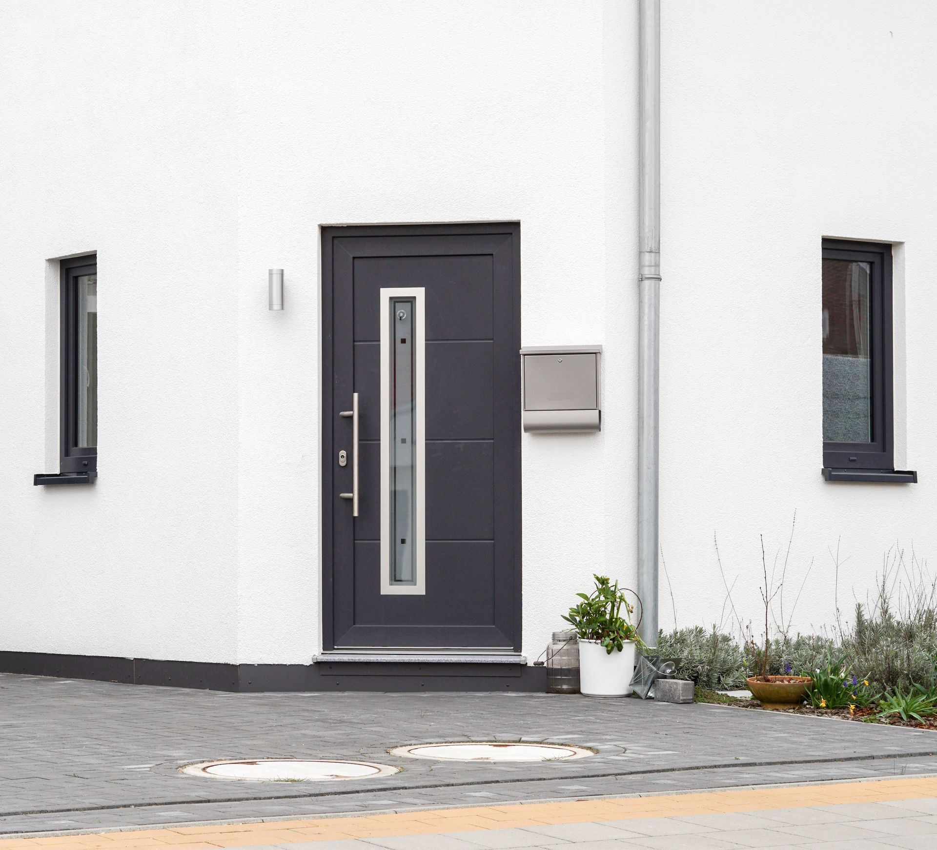 Une porte d'entrée grise avec un panneau de verre vertical sur un mur blanc, flanquée de deux petites fenêtres sombres et d'une allée pavée.