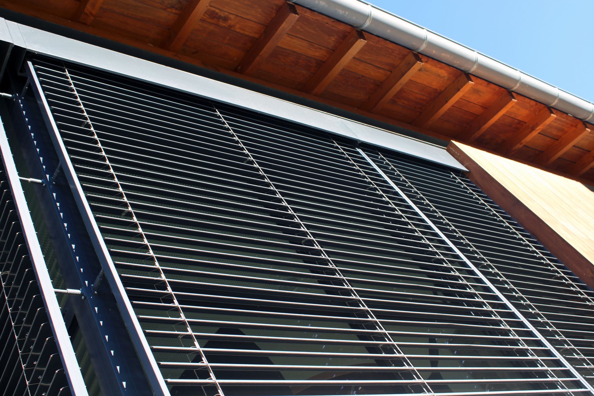 Vue extérieure de stores vénitiens horizontaux en métal noir installés sur une fenêtre d'un bâtiment sous un avant-toit en bois.