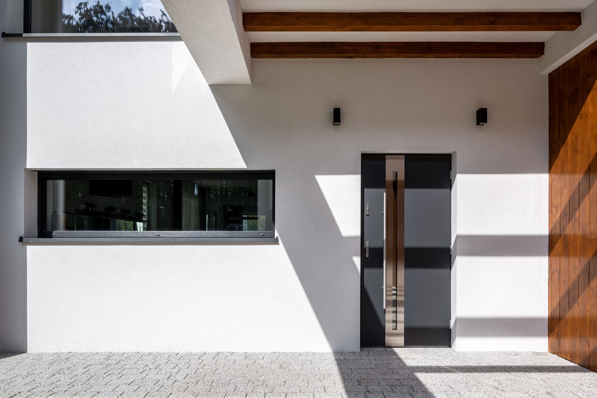 Extérieur de maison moderne avec un mur blanc, une longue fenêtre rectangulaire, une porte sombre avec une poignée en métal et des poutres apparentes au plafond.