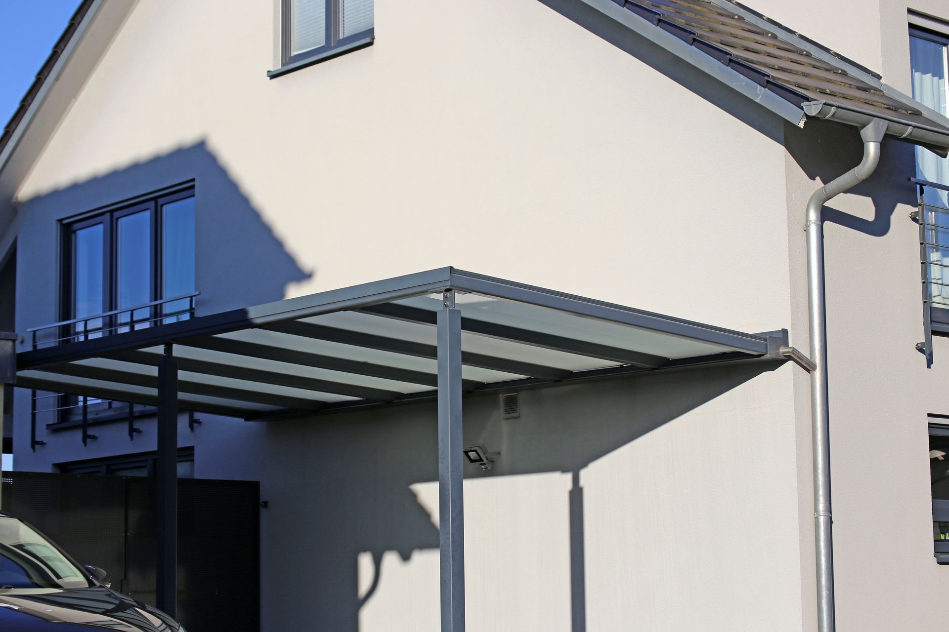 Un abri de voiture moderne en métal gris avec un toit plat translucide, adossé au côté d'une maison d'habitation de couleur claire.