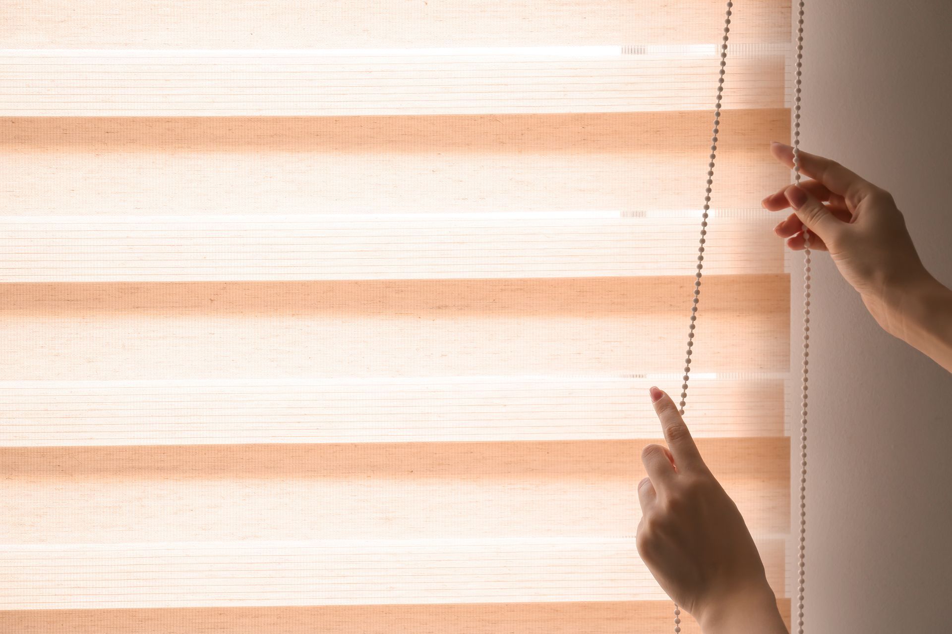 Des mains ajustent une chaînette pour actionner des stores vénitiens à rayures filtrant la lumière.